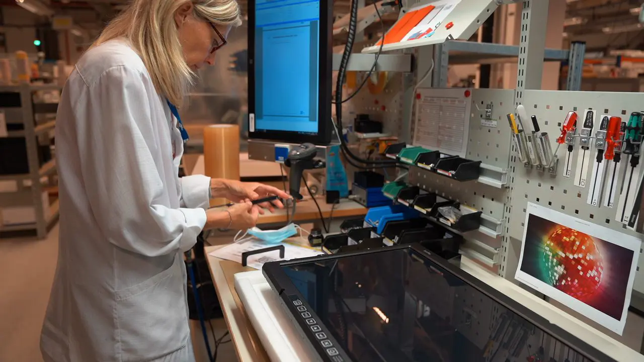"Una operaria en su puesto de trabajo en la planta de ScioTeq, ensamblando un producto electrónico con el apoyo de instrucciones digitales en pantalla."