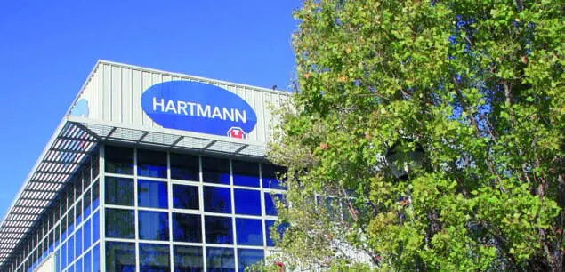 Exterior de un edificio de oficinas moderno con un gran letrero ovalado azul que dice 'HARTMANN', junto a un árbol frondoso y bajo un cielo azul.