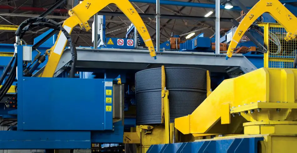 "Maquinaria pesada de color azul y amarillo manipulando una gran bobina de acero corrugado dentro de las instalaciones de producción de CELSA."