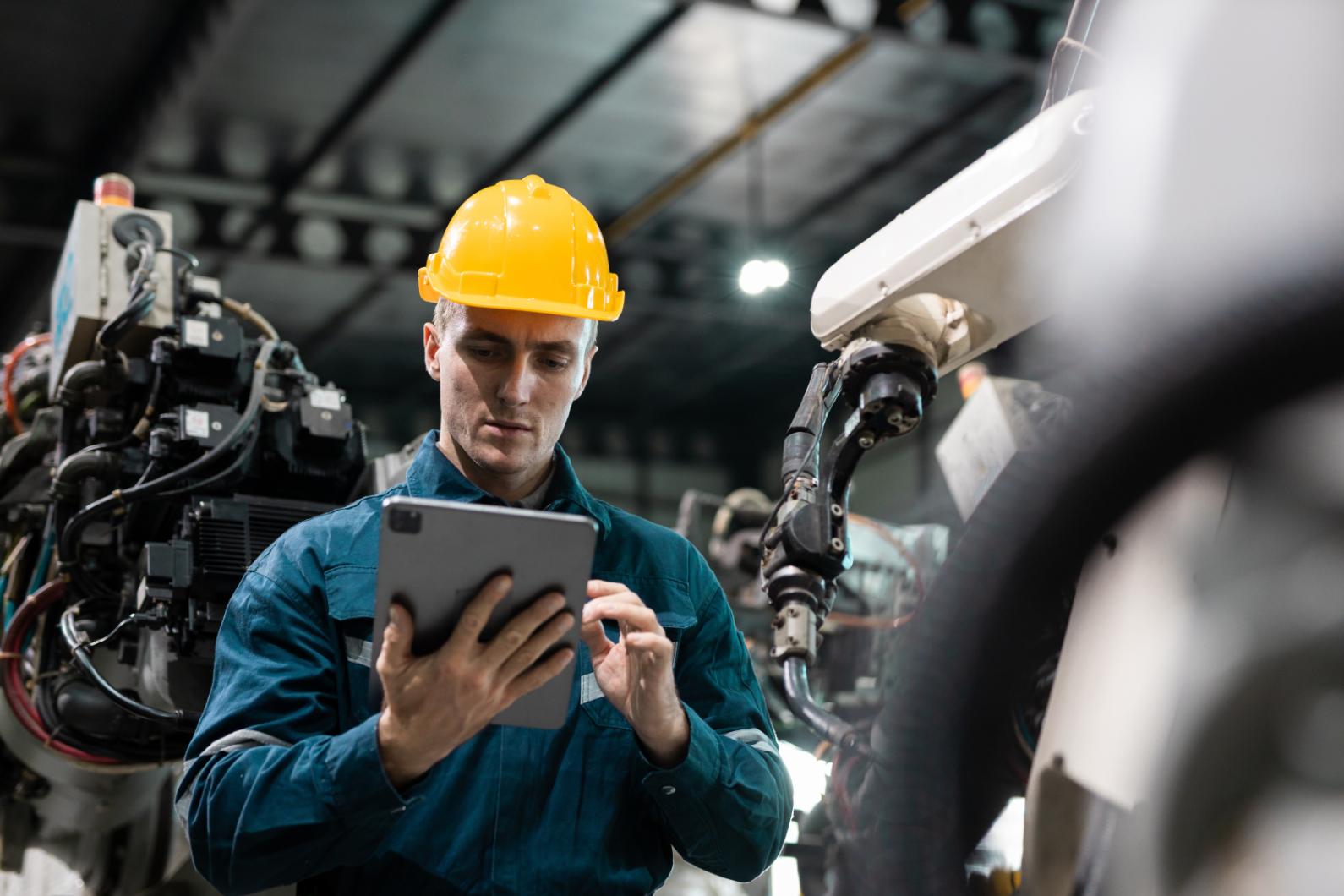 Trabajador con casco de seguridad amarillo utiliza una tableta digital para supervisar un brazo robótico en una fábrica de automatización industrial.
